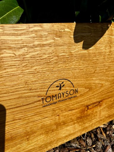 Holzplatte mit eingraviertem Logo "TOMAYSON" und natürlichem Hintergrund.
